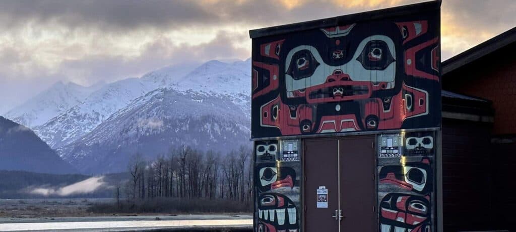 A building with formline art, surrounded by snow-capped mountains.